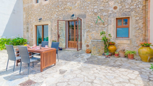 Edificio de piedra con un patio que tiene una mesa y sillas de madera, plantas en macetas y suelo de piedra.