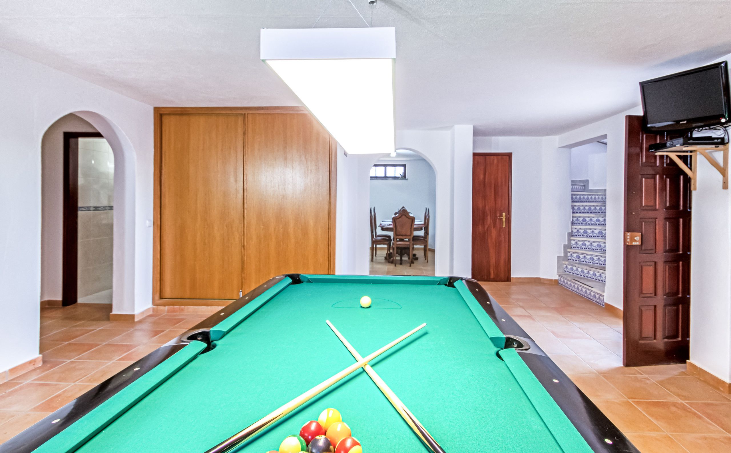 A pool table in a room with a dining area and a TV mounted on the wall.