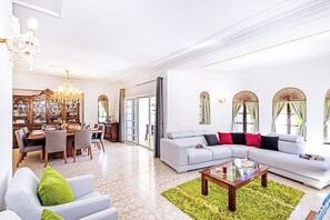 A spacious living room with a large sofa, a dining area with a wooden cabinet, and a chandelier.