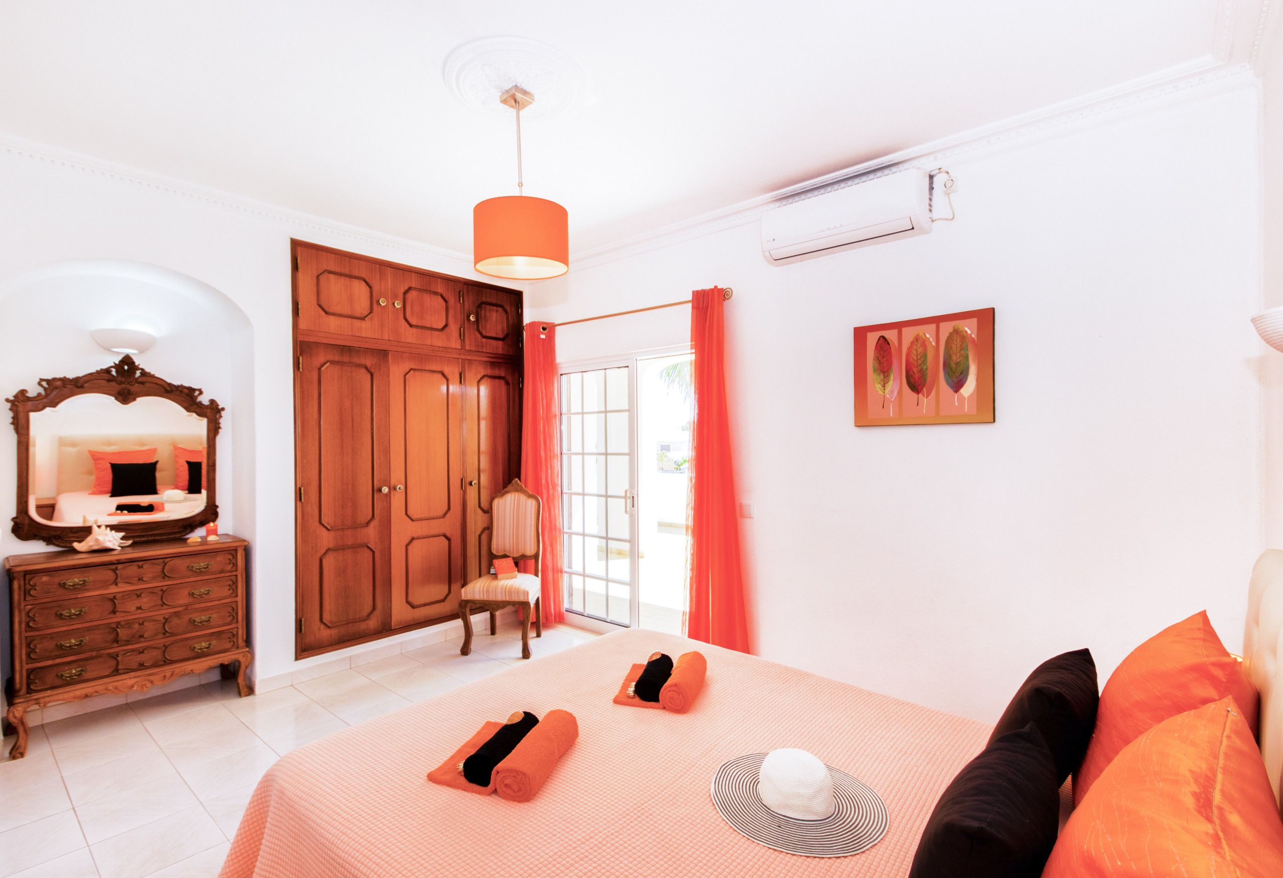 A bedroom with a large wardrobe, a bed with orange bedding, a chair, a mirror, and a picture on the wall.