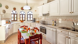 A spacious kitchen with white cabinets, a dining table with chairs, and a large window with arched tops. - Family retreat with heatable pool and BBQ (Albufeira)