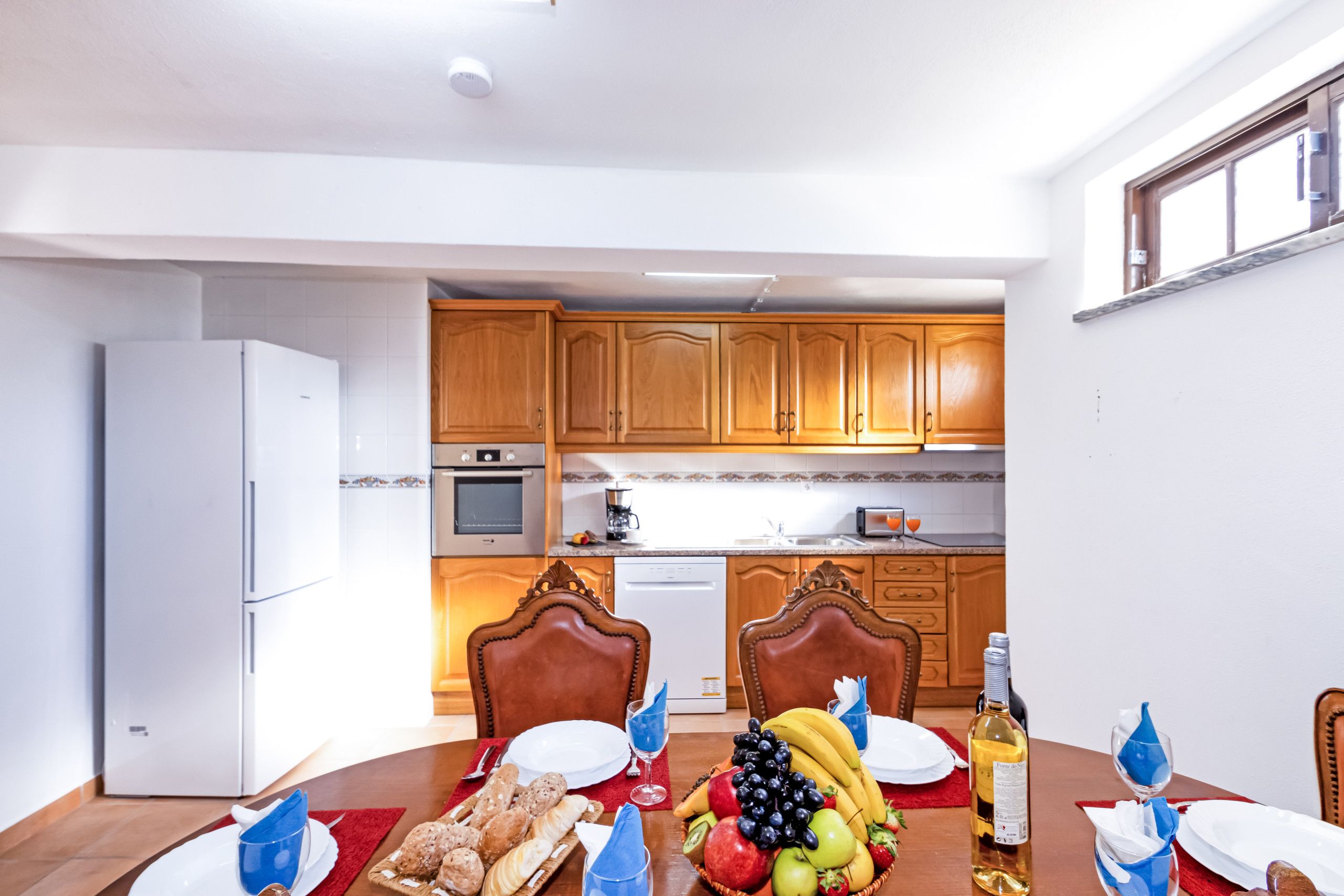 A dining area with a table set for two, a kitchen in the background, and a refrigerator to the left.