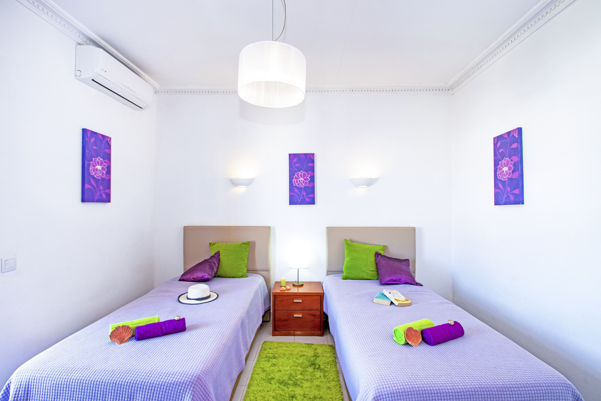 A twin room with two beds, a nightstand, a green rug, and wall-mounted light fixtures.