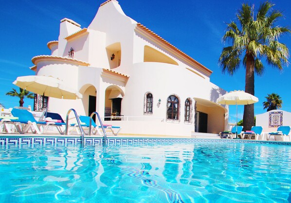 A white building with a swimming pool, palm trees, and sun umbrellas.