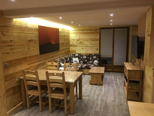 A wooden interior with a dining table, chairs, a sofa, and a television.