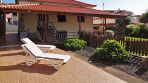 Un bâtiment avec un toit recouvert de tuiles rouges, un patio abrité avec deux chaises longues blanches, et un petit jardin agrémenté de plantes et d’une clôture en bois.