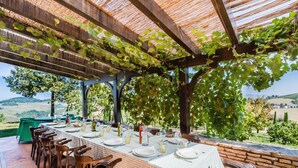 Un espacio comedor al aire libre con una pérgola cubierta de hiedra, una mesa dispuesta para seis personas y vistas al campo.