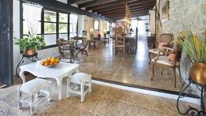 Un vaste espace dédié à la salle à manger, doté d’un haut plafond et de poutres apparentes, comprenant une table blanche sur laquelle est posé un saladier de fruits, plusieurs chaises en bois et un mur en pierre.