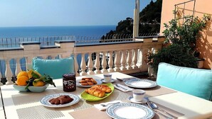 A table set for breakfast with a view of the sea, including plates, cutlery, and a bowl of fruit.