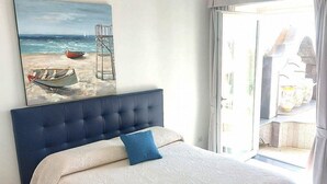 A bedroom with a bed, a painting of a beach scene, and a view of the outside balcony.
