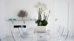 A modern dining area with a glass table surrounded by clear chairs, a white orchid centerpiece, and decorative items on a shelf.