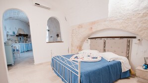 A bedroom with a bed, a stone wall, and a kitchen visible through an archway.