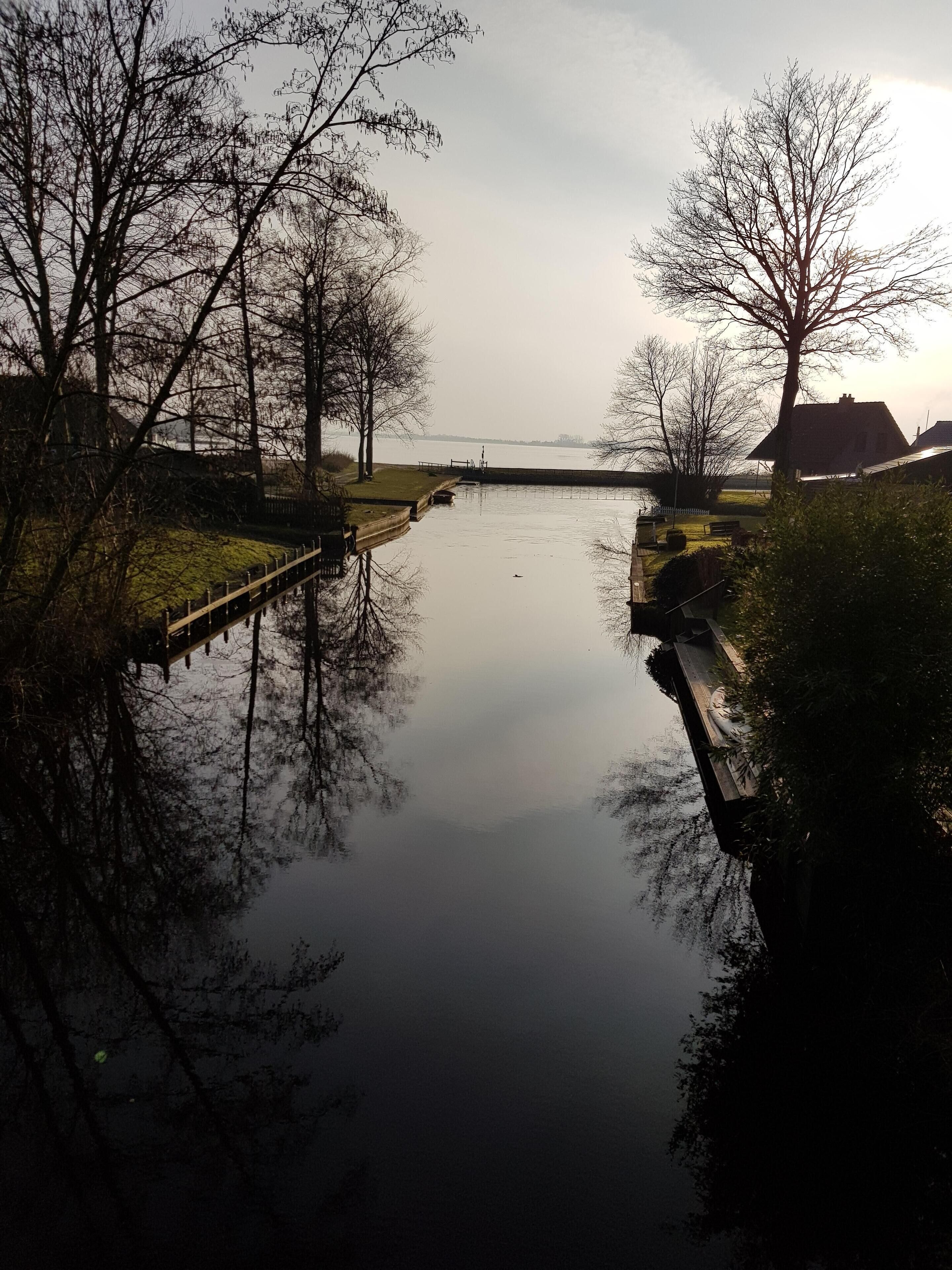A calm river with a wooden bridge, surrounded by leafless trees and a distant view of a house.