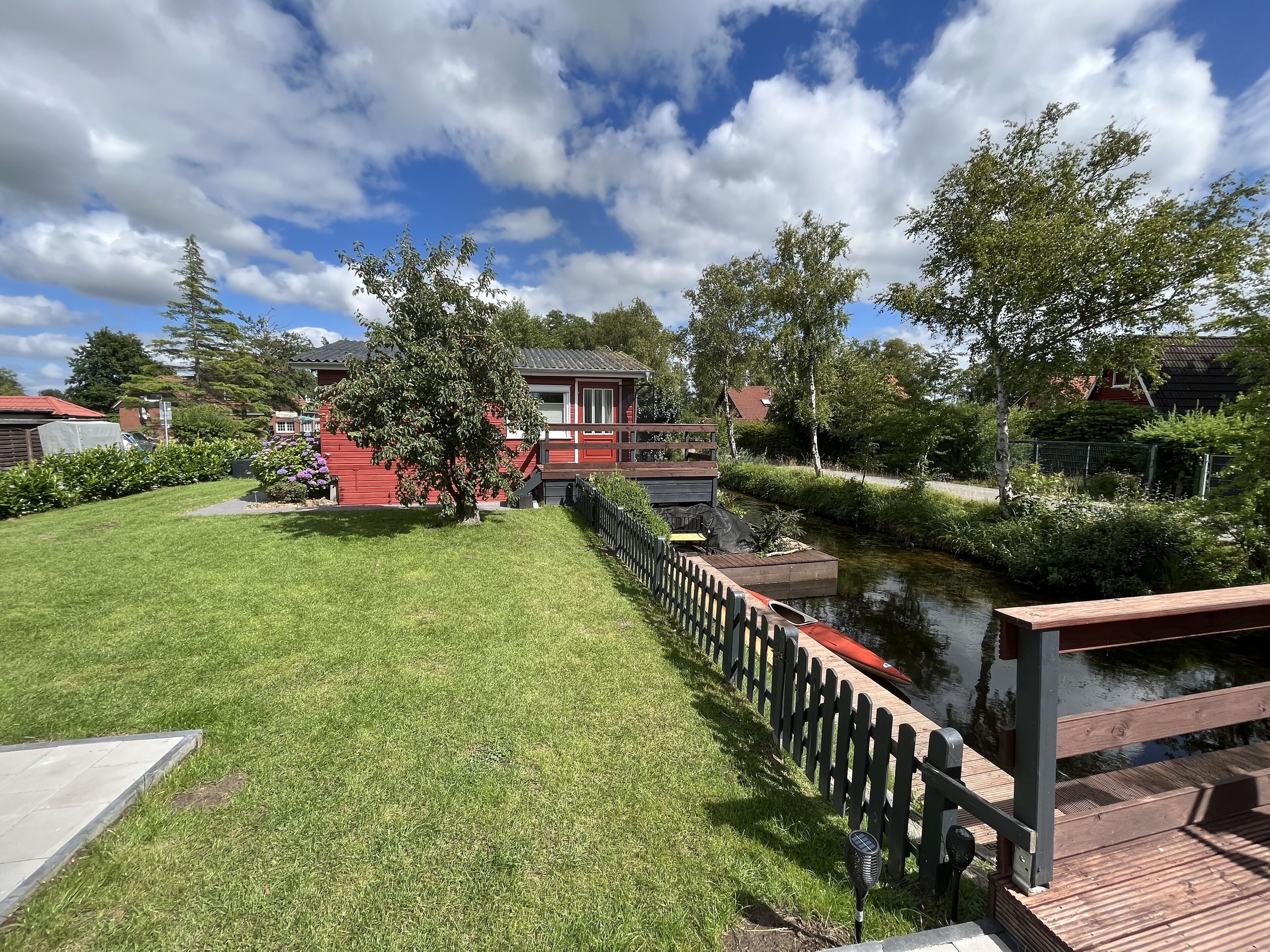 Ein gepflegter Garten mit einer Holterrasse, einem kleinen Teich und einem roten Gebäude im Hintergrund.