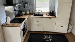 Kompakti keittiö, jossa on valkoiset kaapit, musta työtaso, vedenkeitin, kahvinkeitin ja ikkuna, josta avautuu näkymä vehreään maisemaan.