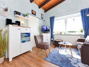 A modern living room with a sofa, a coffee table, a TV, and a kitchen area with a cabinet and shelves. - Apartment "Little Pearl" - Melanie Berres Apartment "Little Pearl" (Esens)