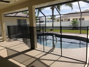 A swimming pool with a metal safety fence, surrounded by a covered patio area with a view of neighboring houses and a lawn. - Last minute deal available (Cape Coral)