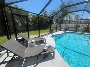 An outdoor swimming pool area with lounge chairs, a poolside table, and a view of the surrounding greenery.