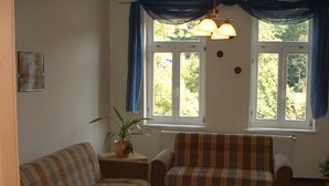 A living room with a sofa, armchair, and a small table with a placemat. There is a window with curtains and a plant on a side table. - Apartment 3 - 3 Bedrooms - 110 m² - Wifi - Dog allowed (Wernigerode)