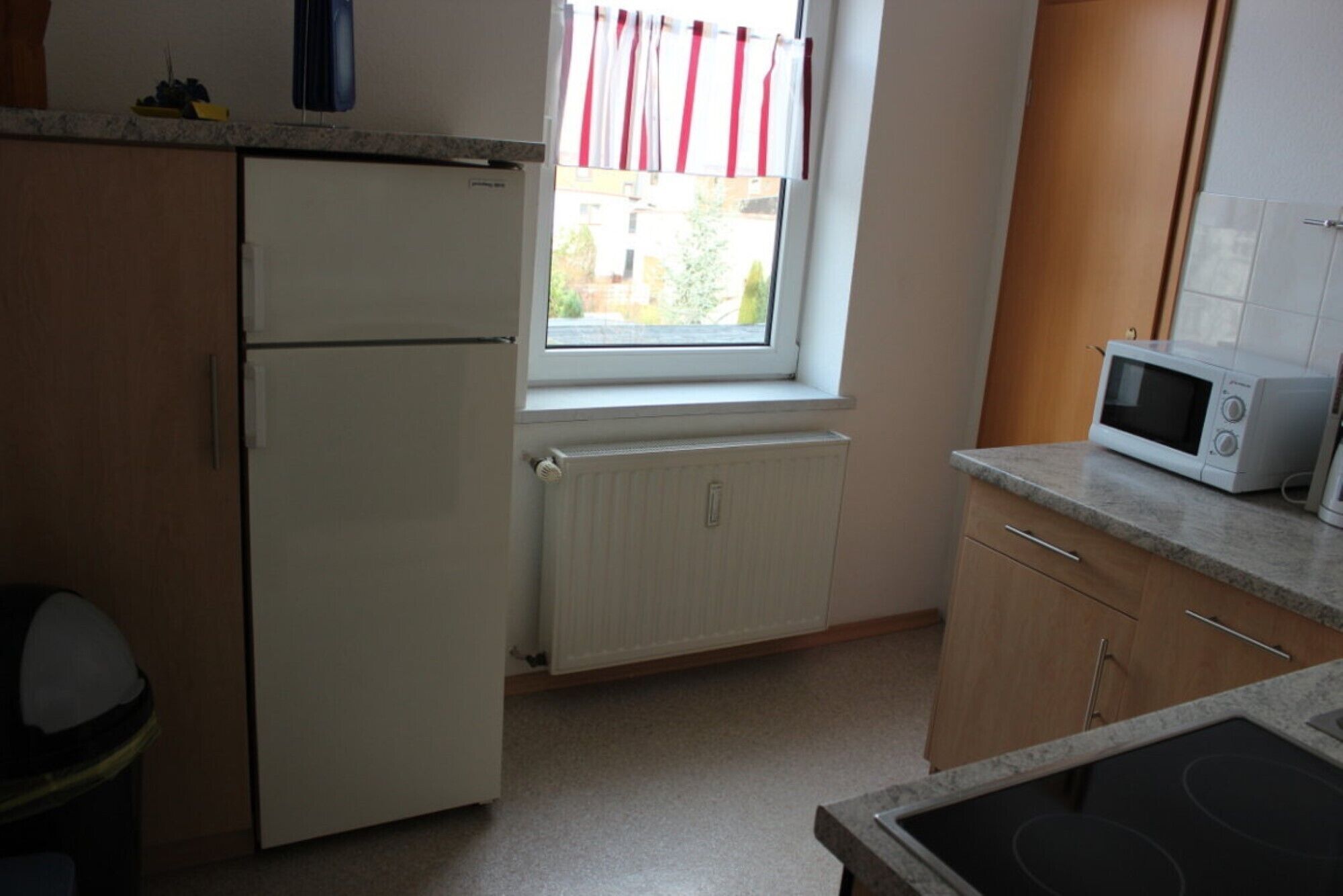 Eine kleine Küche mit Kühlschrank, Mikrowelle, einem Fenster mit Vorhängen und einem Heizkörper.