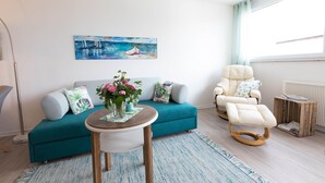 A living room with a teal sofa, a white armchair, a small round table with a vase of flowers, and a painting of sailboats on the wall.