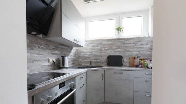 A modern kitchen with grey cabinetry, a built-in oven, a kettle, and a window above the sink.