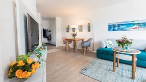 A modern living room with a teal sofa, a coffee table, a dining area with a wooden table and chairs, and a TV mounted on the wall.