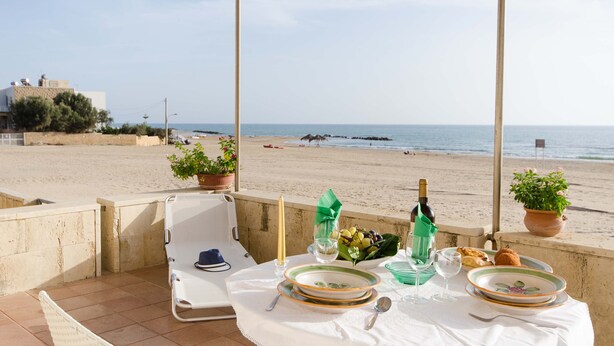 A table set for two on a terrace overlooking a beach, with a view of the sea and a building in the background. - Small, modern beach house on the sand, quiet,sea view, WiFi, aircon, max. 7 p. (Donnalucata)