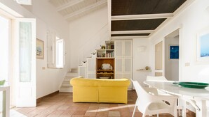 A bright, open living area with a yellow sofa, white chairs, and a dining table. A staircase with bookshelves leads to an upper level. - Small, modern beach house on the sand, quiet,sea view, WiFi, aircon, max. 7 p. (Donnalucata)
