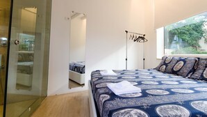A bedroom with a glass shower enclosure, a large mirror, a bed with patterned bedding, and a view of greenery outside. - Small, modern beach house on the sand, quiet,sea view, WiFi, aircon, max. 7 p. (Donnalucata)