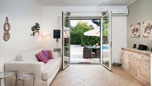 A modern living room with a sofa, two glass tables, a cabinet, and a sliding glass door leading to an outdoor area with a pool and umbrellas. - Monte - Independent House with private Pool, 4 People, Garden WiFi, A/C, Sicily (Ragalna)