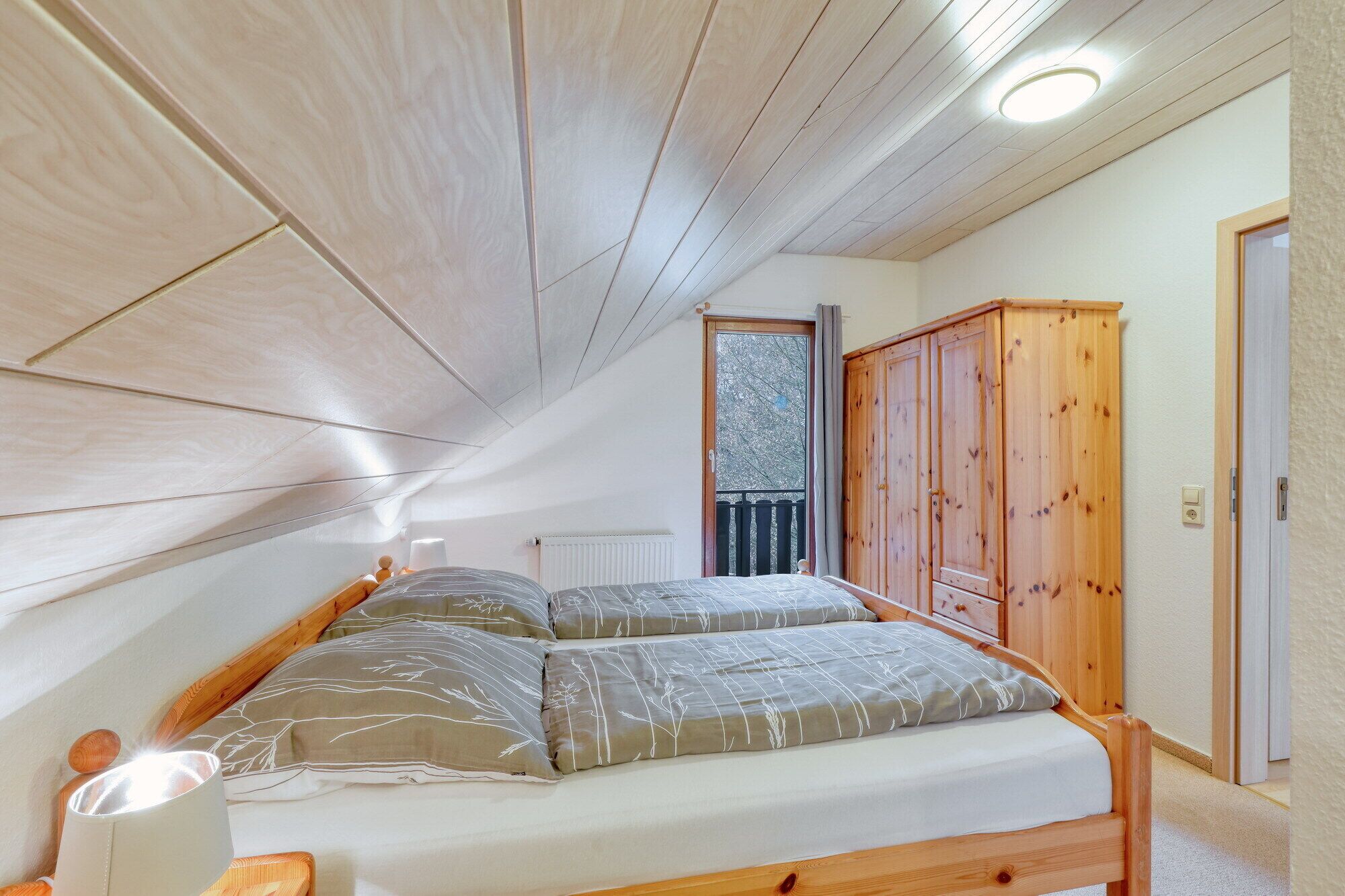 A bedroom with a sloped ceiling, wooden ceiling beams, a bed with grey bedding, a wooden wardrobe, and a view of a balcony with a railing.