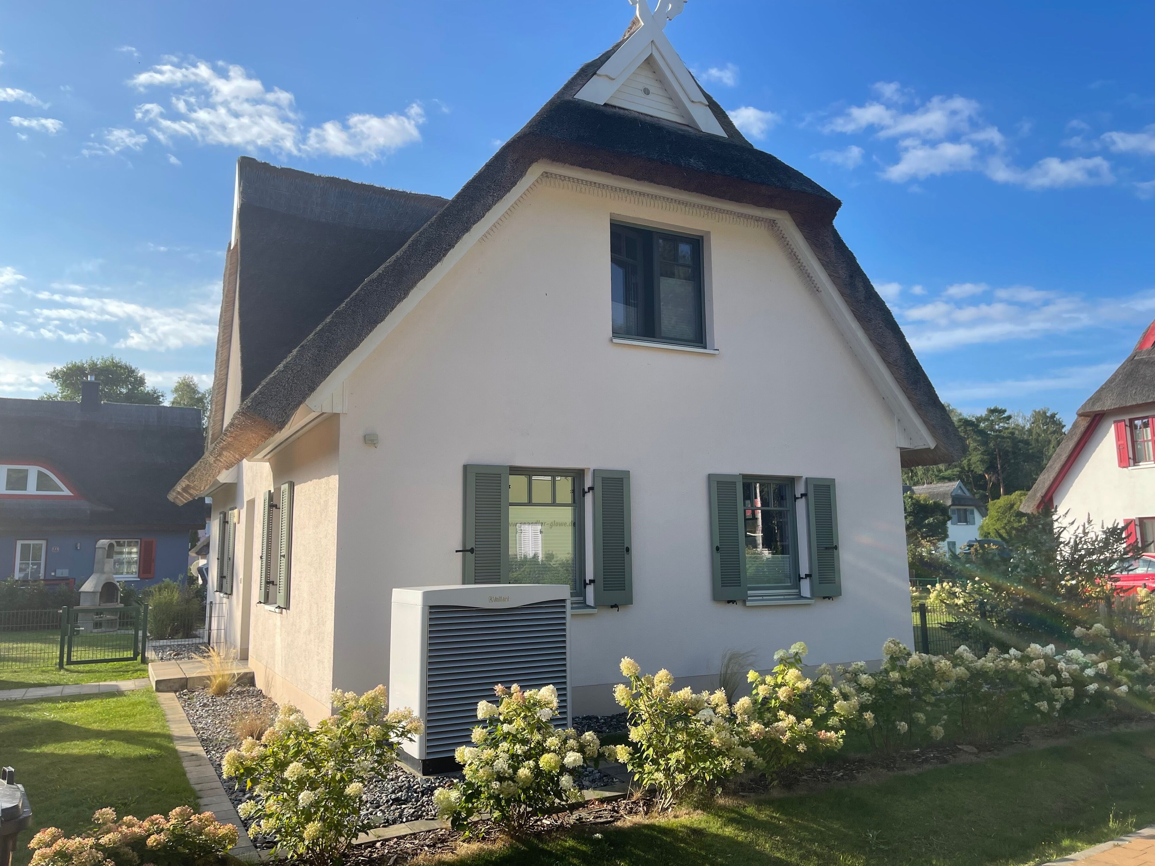 Ein weißes Haus mit grünen Fensterläden, einem geneigten Dach und einem kleinen Garten mit weißen Blumen.