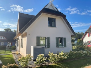 Une maison blanche avec des volets verts, un toit en pente et un petit jardin où poussent des fleurs blanches.