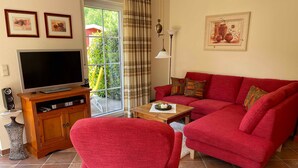A living room with a red sofa, a TV on a wooden stand, a table with a candle, and a large window with curtains. - Schüßler house The red house - Schüßler (Trassenheide)
