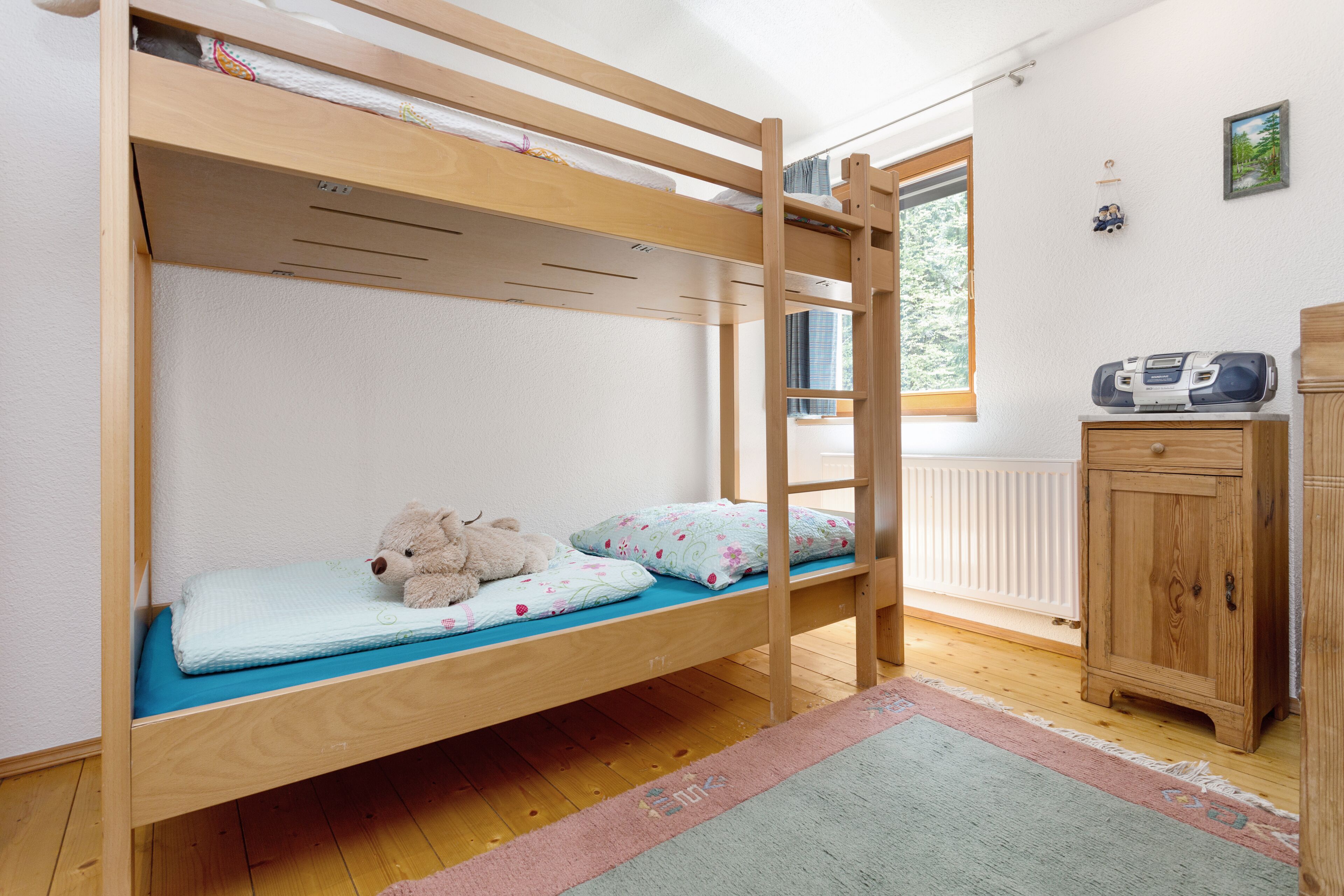 Un lit superposé avec un ours en peluche et des oreillers, une armoire en bois et une fenêtre donnant sur les arbres.