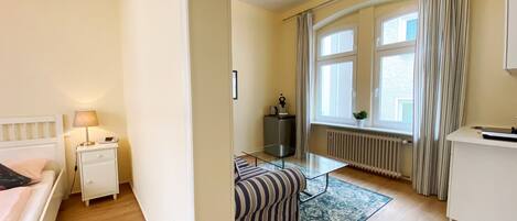 A hotel room with a bed, bedside table, lamp, radiator, window with curtains, and a small table with a plant.