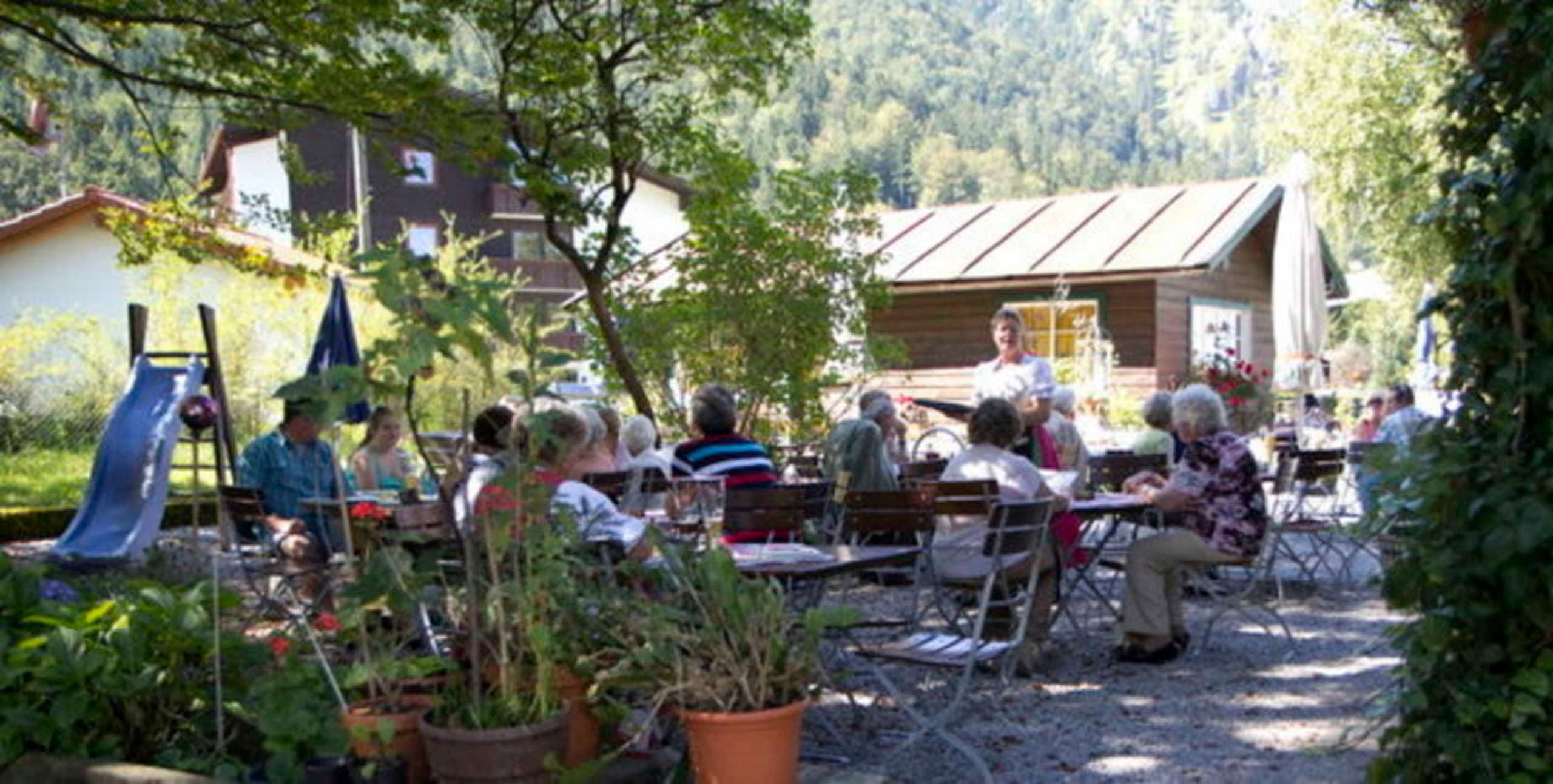Un espace extérieur aménagé avec des tables et des chaises, entouré de plantes en pot et d’arbres.