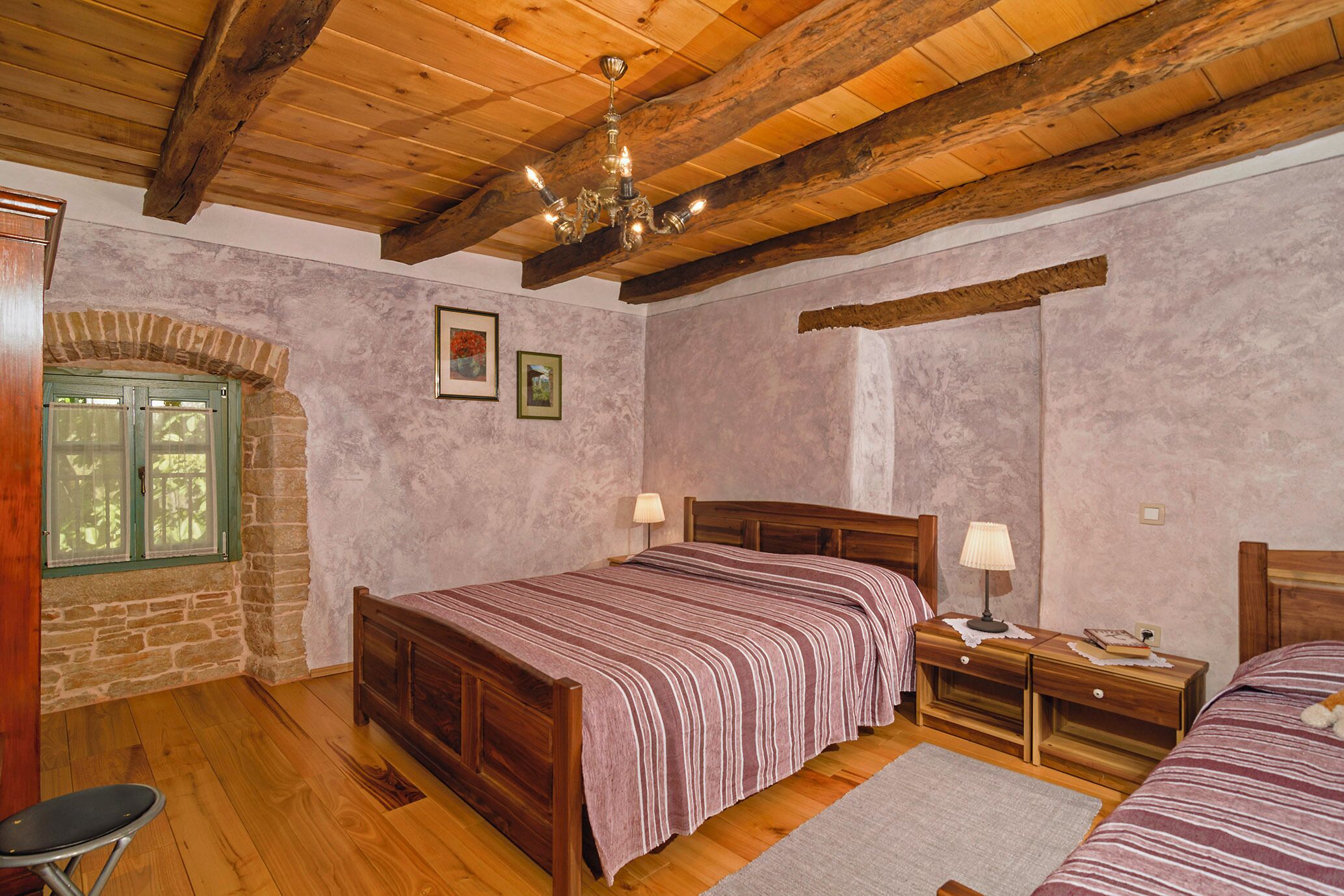 A bedroom with a wooden ceiling, stone walls, a bed with a striped bedspread, two bedside tables with lamps, and a small chair.