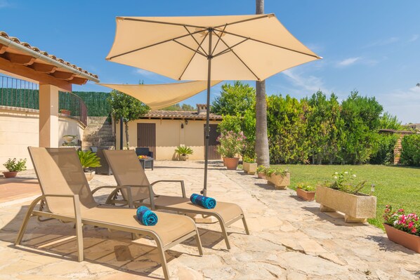 A patio with two beige lounge chairs, a large beige umbrella, and potted plants.
