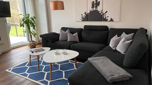 A modern living room with a dark grey sofa, two white side tables, a blue rug, and a large window with a view of greenery.