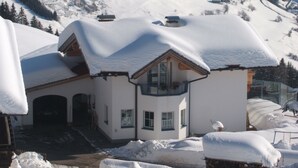 Un chalé cubierto de nieve con un balcón, una escalera de madera y vistas del paisaje nevado circundante.