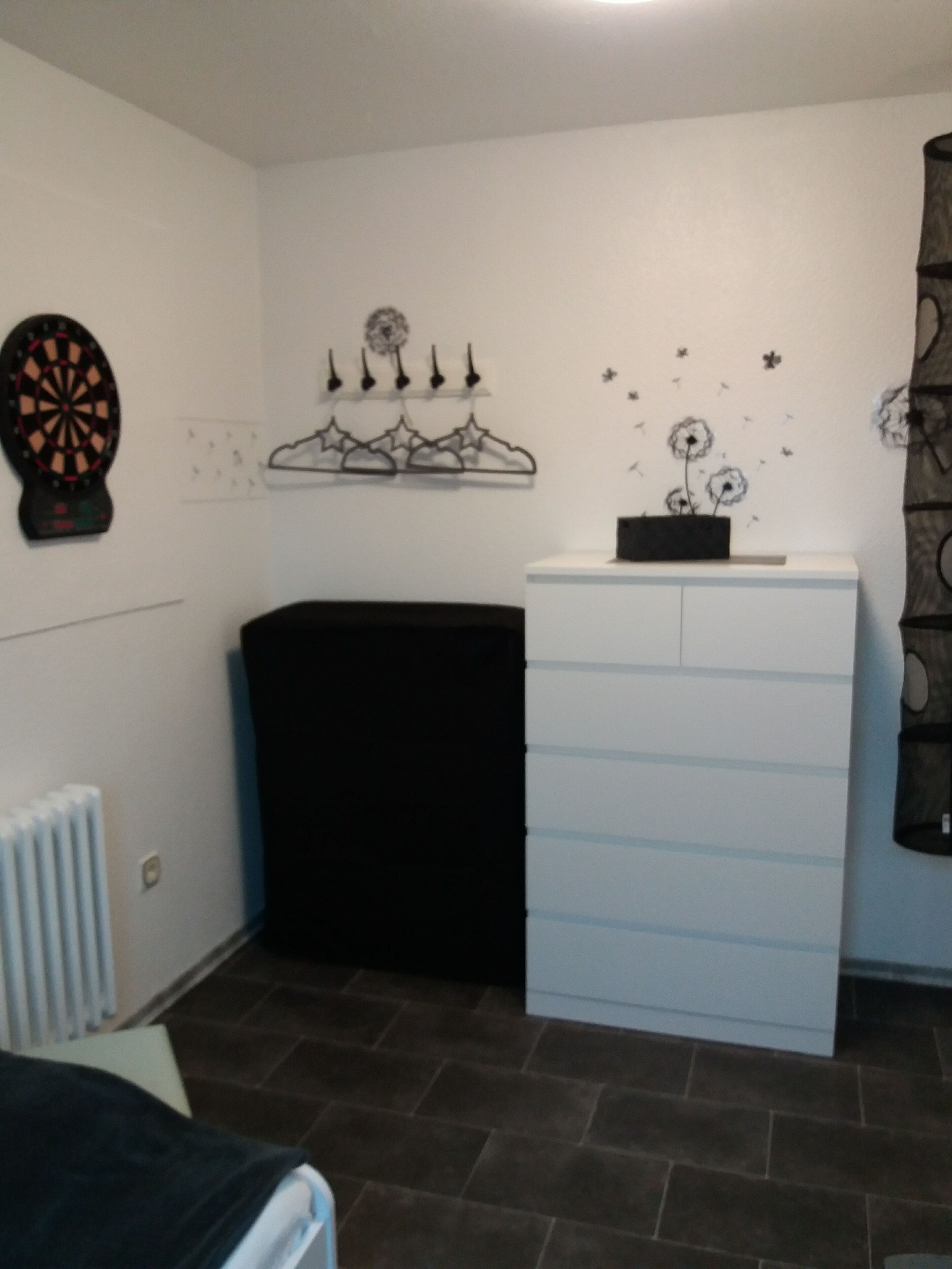 Une pièce comprenant un lit, une commode blanche, un pouf noir, un jeu de fléchettes et des éléments décoratifs sur les murs, notamment des motifs floraux et en forme d’étoile.