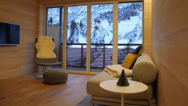 L’intérieur chaleureux d’un chalet avec un plancher en bois, un fauteuil rembourré, un canapé avec des coussins, une petite table ronde et une grande fenêtre donnant sur un paysage montagneux enneigé.