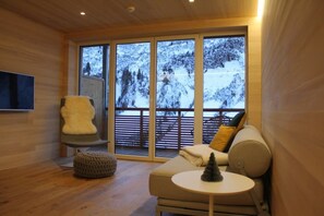 Interior acogedor de una cabaña con suelo de madera, un sillón mullido, un sofá con cojines, una mesita redonda y un amplio ventanal con vistas a una montaña nevada.