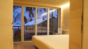 Une chambre avec un plancher en bois, un lit, une télévision et une porte-fenêtre coulissante donnant sur un balcon avec vue sur un paysage enneigé.
