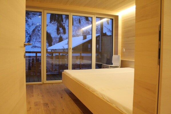 Un dormitorio con piso de madera, cama, televisor y puerta de vidrio corredera que da a un balcón con vistas a un paisaje nevado.