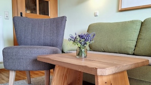 A room with a wooden table, a grey armchair, a green sofa, a wooden shelf with a lamp, and a framed picture on the wall. - Heissbauer Vacation Rental "Karwendelblick" with Mountain Views, 2 Bedrooms, and 2 Bathrooms (Wackersberg)