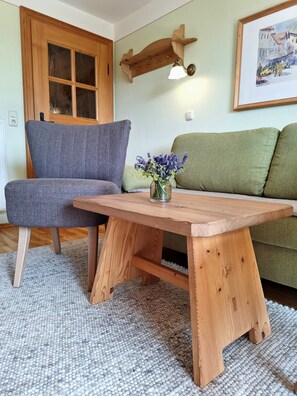 Una habitación con una mesa de madera, un sillón gris, un sofá verde, una estantería de madera con una lámpara y un cuadro en la pared.