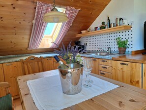 Una cocina de madera con una mesa de comedor, una lámpara colgante, una ventana con cortinas y una estantería con utensilios de cocina.